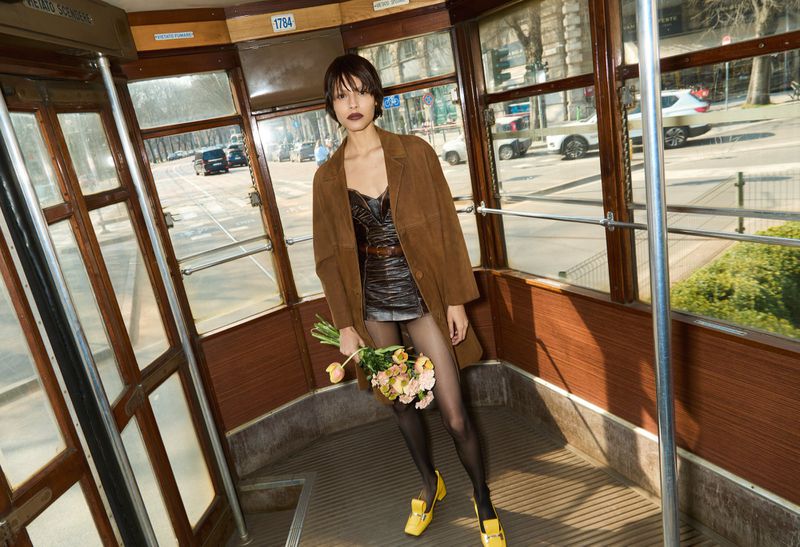 Woman in brown coat poses inside tram with flowers, wearing yellow shoes, black stockings, and a dress. Urban transport scene with natural light.