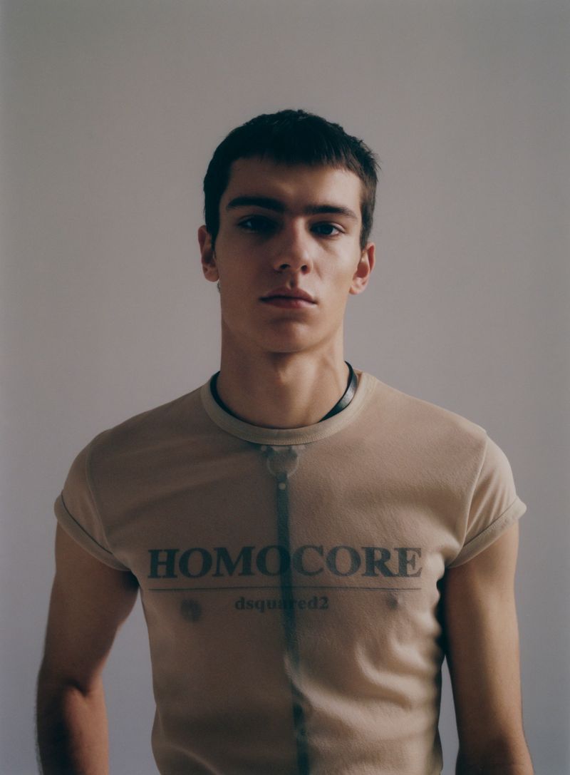 Portrait of a young man in a tan t-shirt with 'HOMOCORE dsquared2' printed on the front, posing against a plain neutral background.