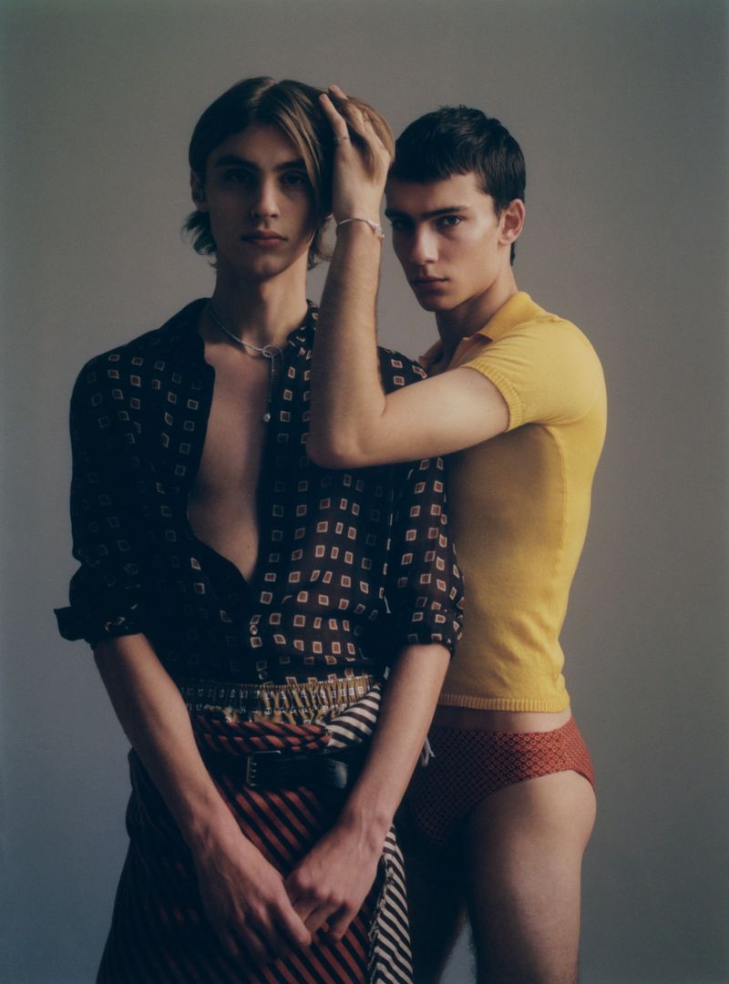 Two young men pose in a photo studio. One man wears an open patterned shirt, a striped wrap, and a necklace. The other wears a yellow polo shirt.