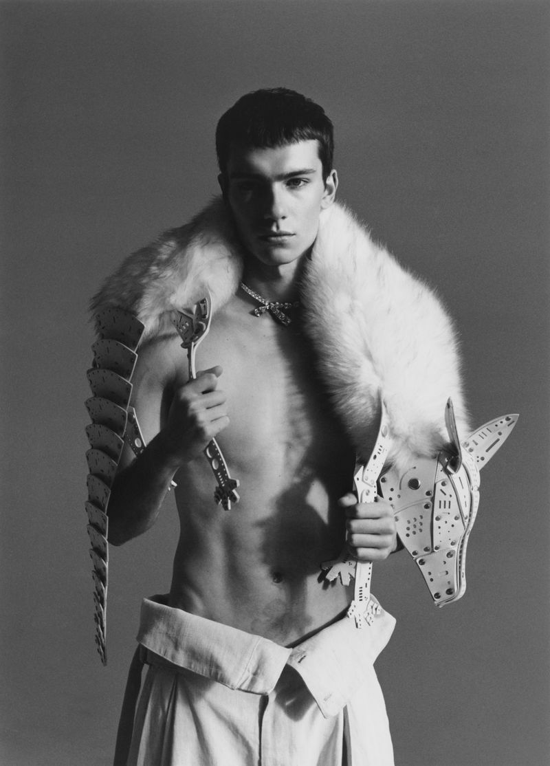 A shirtless male model poses indoors in a studio wearing a fur shawl and avant-garde clothing and holding unique metal accessories.