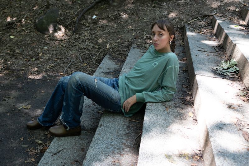 A young adult female with long brown hair is sitting on concrete steps outdoors wearing a long-sleeved shirt, jeans and brown shoes.