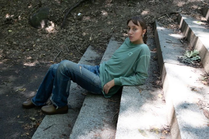 A young adult female with long brown hair is sitting on concrete steps outdoors wearing a long-sleeved shirt, jeans and brown shoes.