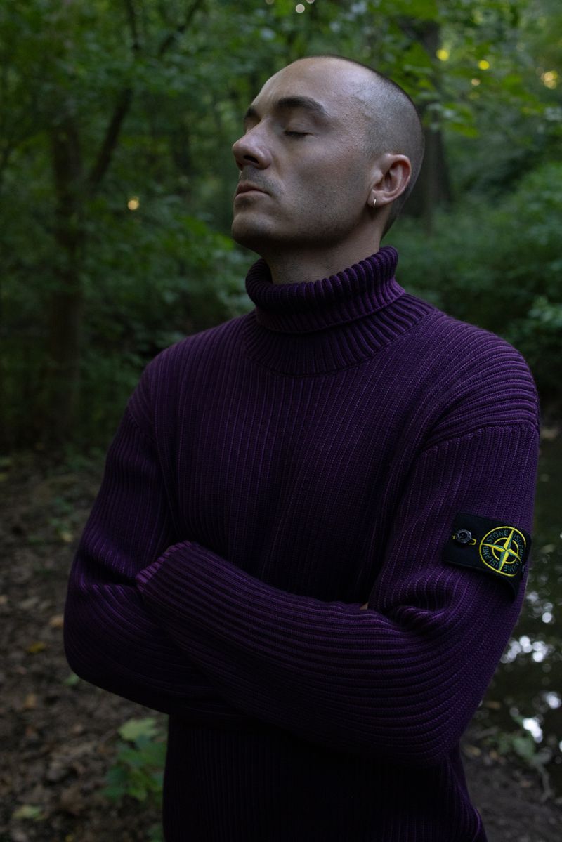 Man in purple turtleneck sweater stands with arms crossed in a forest, eyes closed, looking serene. Natural light, outdoor portrait.