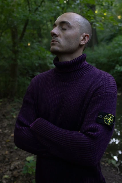 Man in purple turtleneck sweater stands with arms crossed in a forest, eyes closed, looking serene. Natural light, outdoor portrait.
