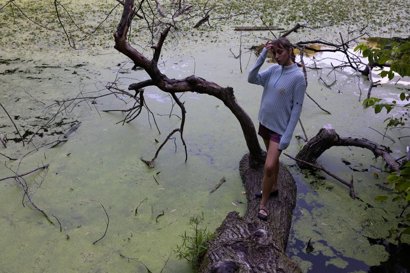 Young woman standing on a fallen tree trunk over a pond with green algae in an outdoor, summertime setting with natural lighting.