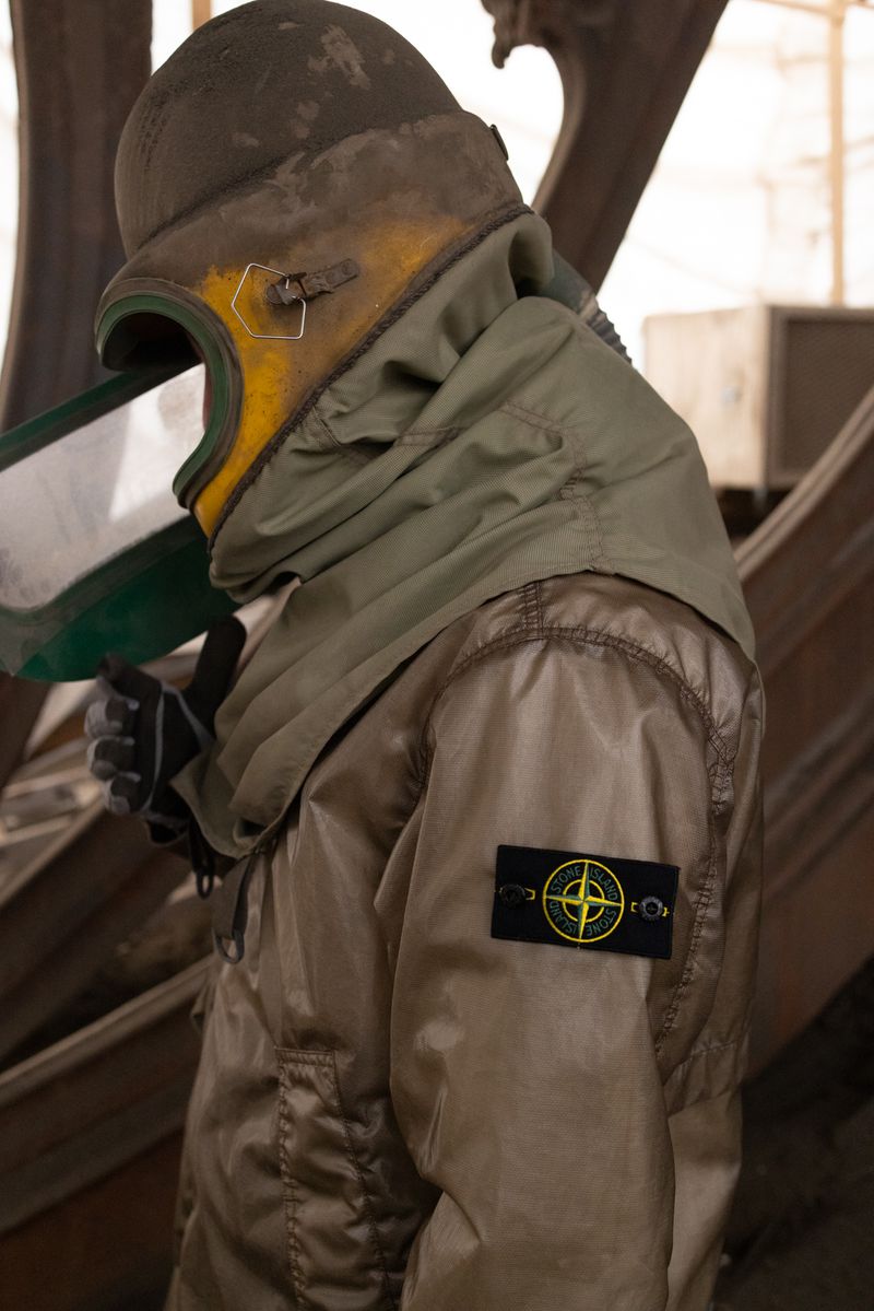 Person in protective suit with hooded jacket featuring a Stone Island logo, standing against a rough, industrial background.