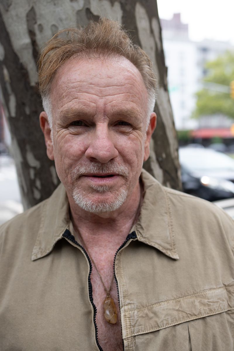 Portrait of a mature adult man with short red hair wearing a tan jacket and a necklace standing outdoors on a city street.
