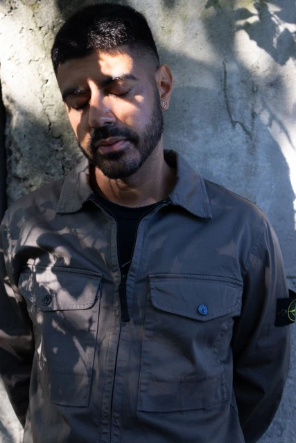 Portrait of a man with dark hair and beard, eyes closed, wearing a grey shirt, against a textured wall with tree shadows.