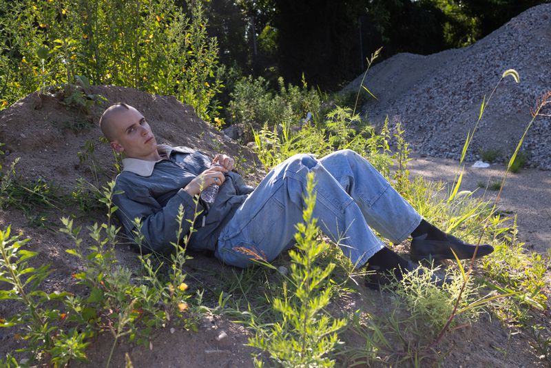 Young adult male poses on a grassy hill in natural light wearing a blue denim outfit and holding a water bottle in an outdoor setting.