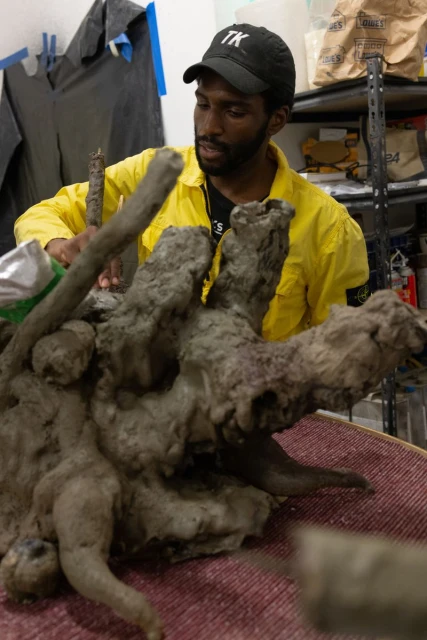 Adult male artist working on a grey sculpture in a bright yellow jacket, indoors. The artwork is displayed on a maroon cloth.