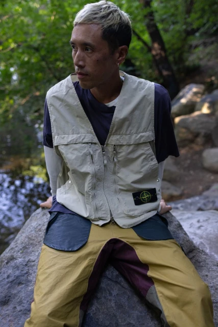Man in nylon vest and color-blocked pants poses on a rock in a forest setting with dappled sunlight, looking off to the side.