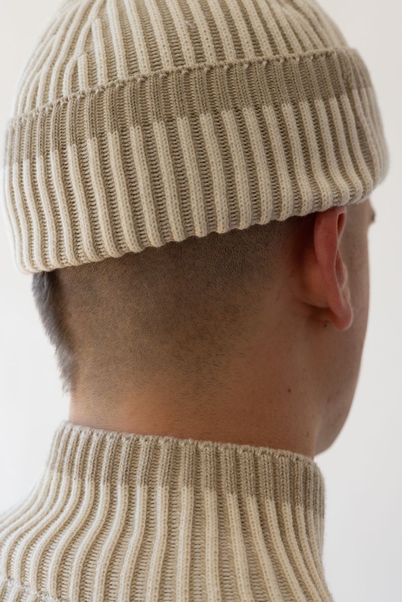 Close-up of a person wearing a cream-colored, ribbed knit hat and turtleneck sweater, facing away from the camera in a neutral setting.