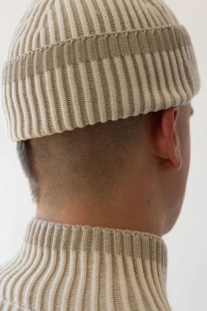 Close-up of a person wearing a cream-colored, ribbed knit hat and turtleneck sweater, facing away from the camera in a neutral setting.