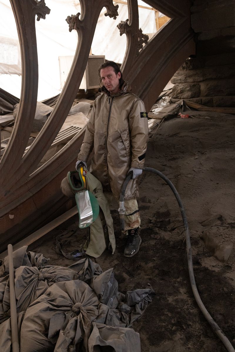 Adult male in coveralls stands holding a sandblasting tool in a stone building with detailed window framing in a low angle shot.