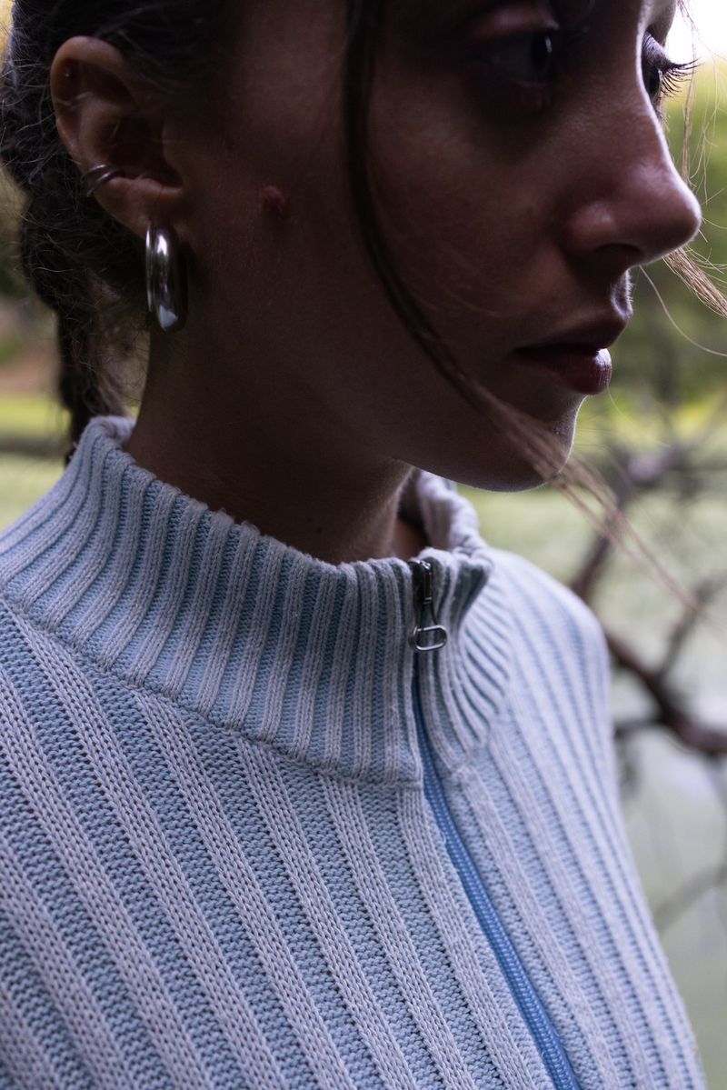 Close-up of a young woman with silver hoop earrings wearing a light blue, ribbed, zip-up sweater, with a blurred outdoor background.