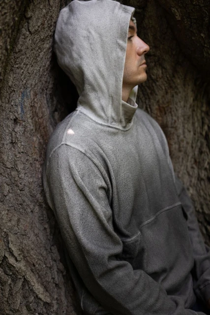 Portrait of a young adult man wearing a hoodie, leaning against a tree, gazing off to the side in an outdoor, natural setting.
