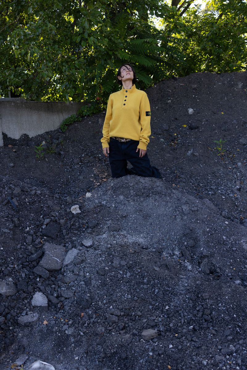 Person in a yellow sweater kneeling on a rocky hill outdoors with a tree in the background, gazing upwards toward the sky.
