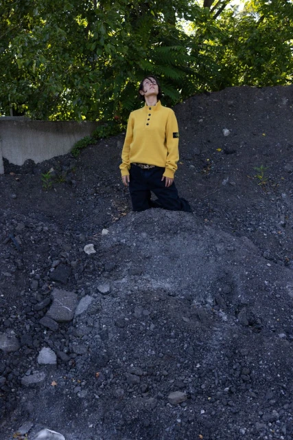 Person in a yellow sweater kneeling on a rocky hill outdoors with a tree in the background, gazing upwards toward the sky.