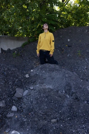Person in a yellow sweater kneeling on a rocky hill outdoors with a tree in the background, gazing upwards toward the sky.