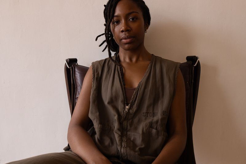 A young Black woman with dreadlocks sits in a brown leather chair wearing a sleeveless olive green jumpsuit against a cream wall.