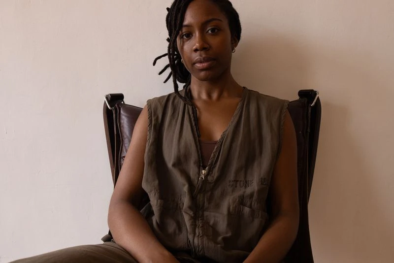 A young Black woman with dreadlocks sits in a brown leather chair wearing a sleeveless olive green jumpsuit against a cream wall.