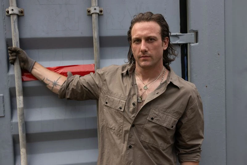 Adult male poses near a grey shipping container, wearing a brown work shirt, multiple necklaces, and gloves while gripping a metal bar.