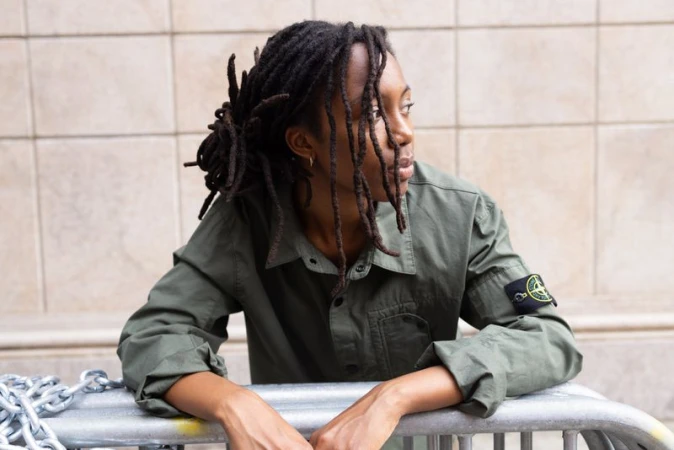 Young woman with dreadlocks in olive green shirt standing behind a metal barricade in an outdoor setting, looking over her shoulder.