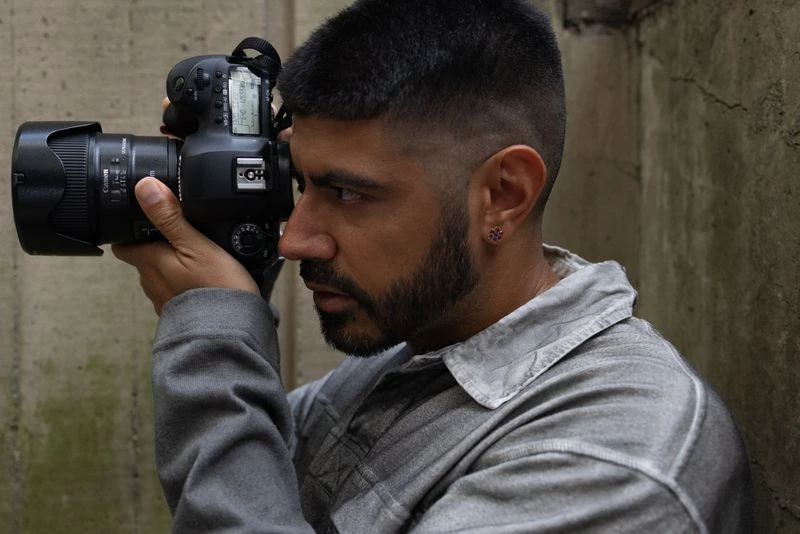 Adult male photographer holding a DSLR camera at eye-level, wearing a grey long-sleeve shirt, with a concrete wall as the background.