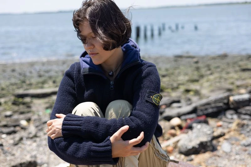A young woman with short brown hair sits on a rocky beach, hugging her knees while wearing a blue sweater and tan pants on a sunny day.