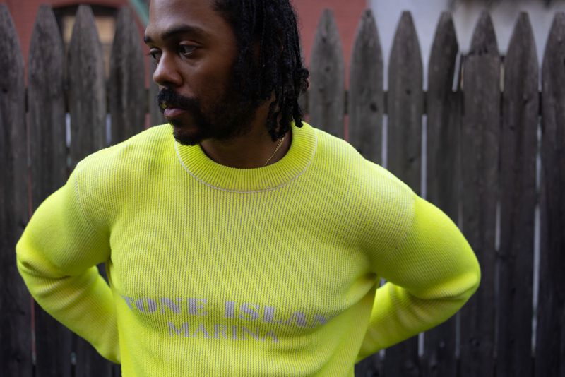 Portrait of a handsome adult male with dark hair, wearing a bright yellow sweater, posing against a wooden fence in an outdoor setting.