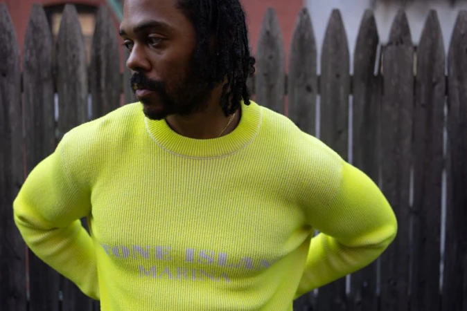 Portrait of a handsome adult male with dark hair, wearing a bright yellow sweater, posing against a wooden fence in an outdoor setting.