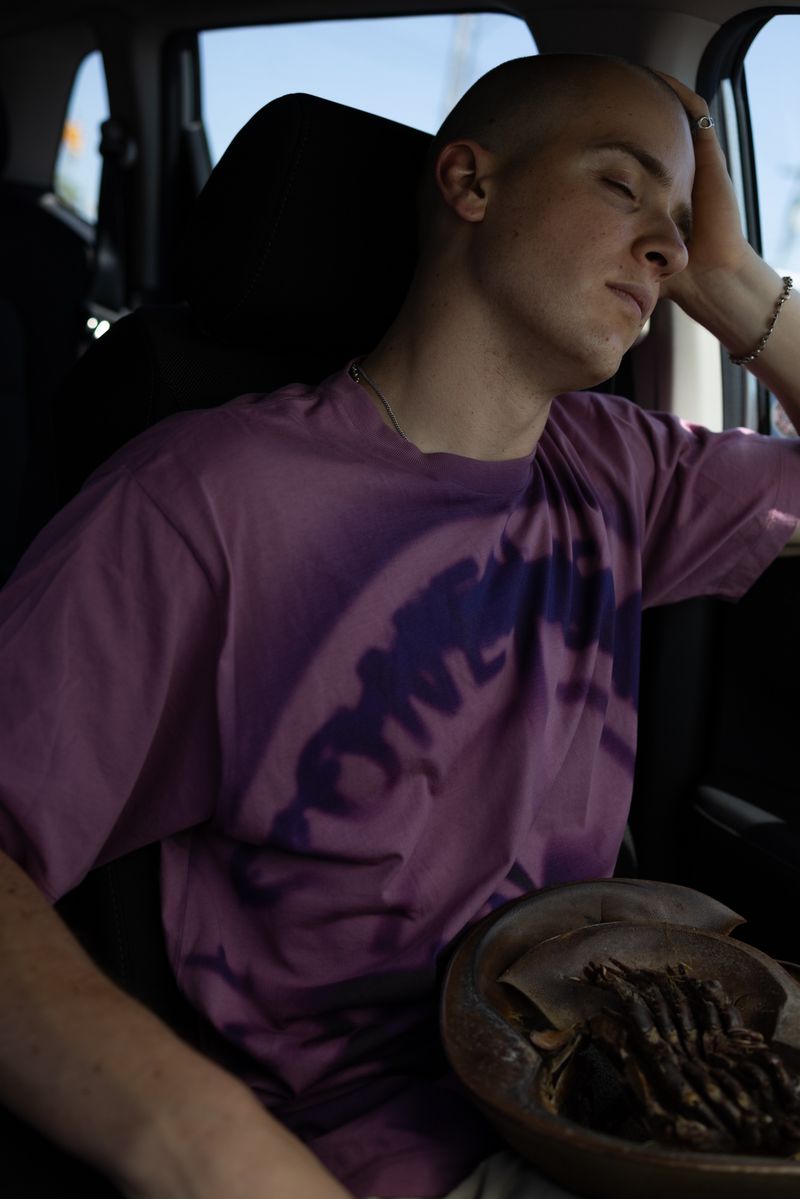 Adult male sits in car with head resting on hand; he is wearing a purple t-shirt; a bowl with food rests in his lap.