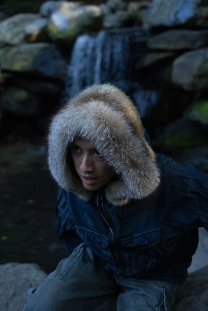 An adult male poses near a small waterfall while wearing a blue hooded parka with fur trim in an outdoor setting.