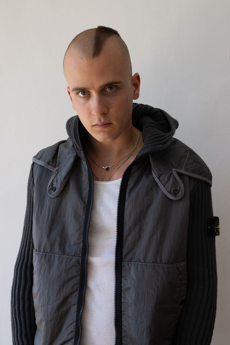 Man with a mohawk and gray hooded jacket poses against a neutral background, looking directly at the camera in a portrait-style shot.
