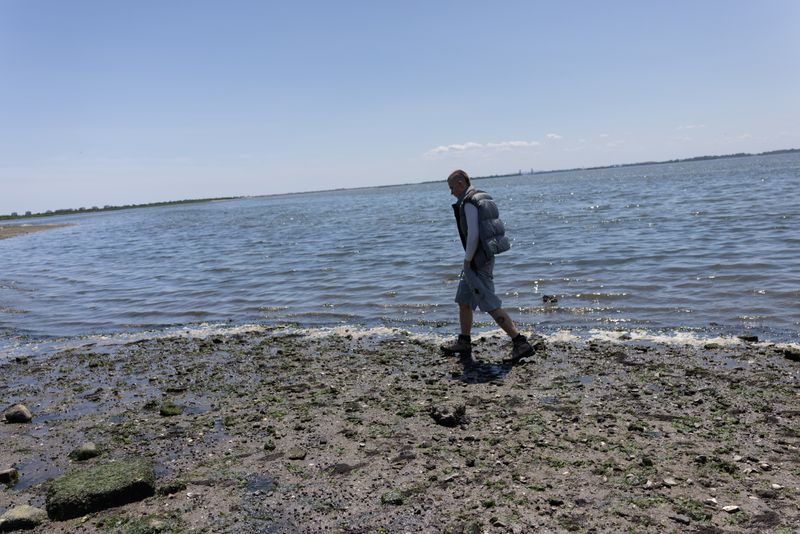 Adult male walking on the shore near the water with a puffy vest and shorts on a sunny day near the ocean, in a medium shot.