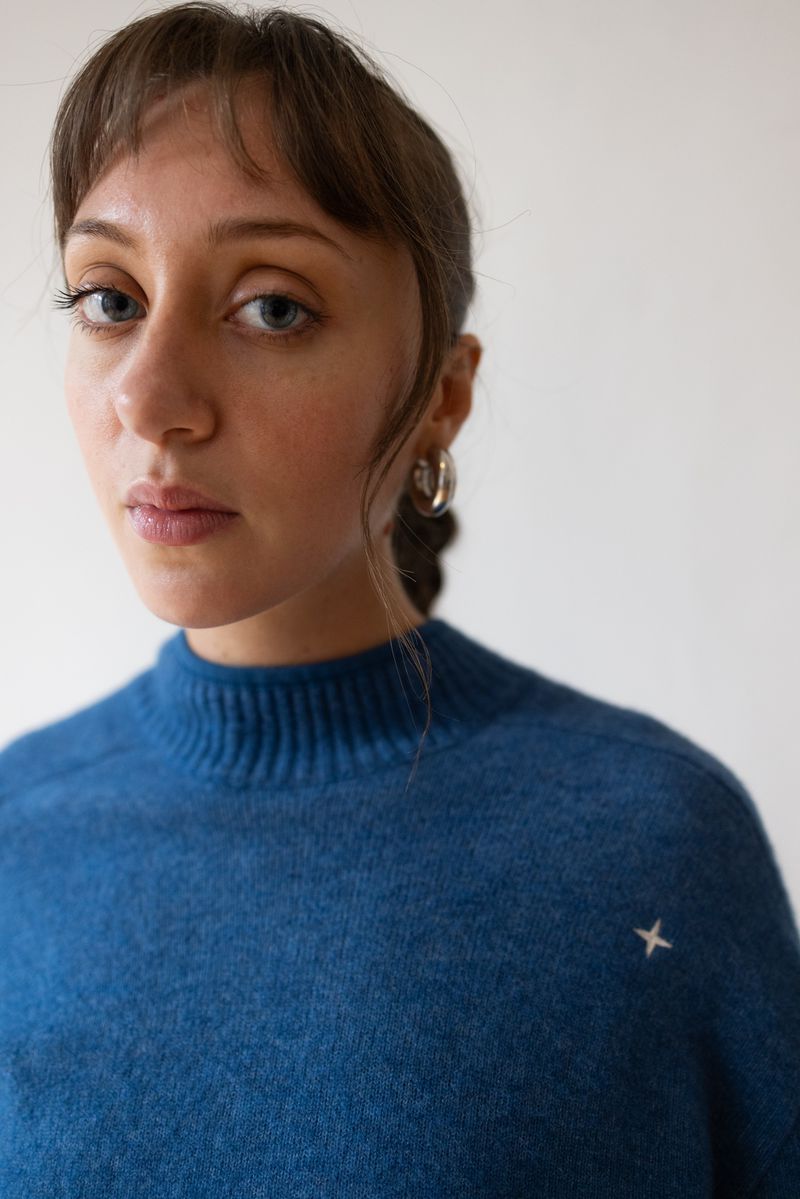 Close-up portrait of a young woman with light skin and blue eyes wearing a blue sweater against a plain white background in a photo studio.