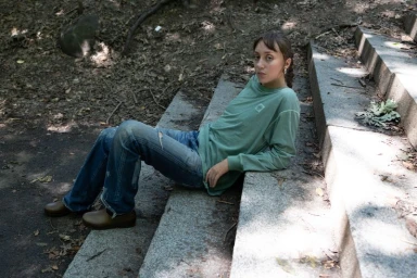 Model wearing a sage green long-sleeve top and blue denim jeans seated on stone outdoor steps outdoors.