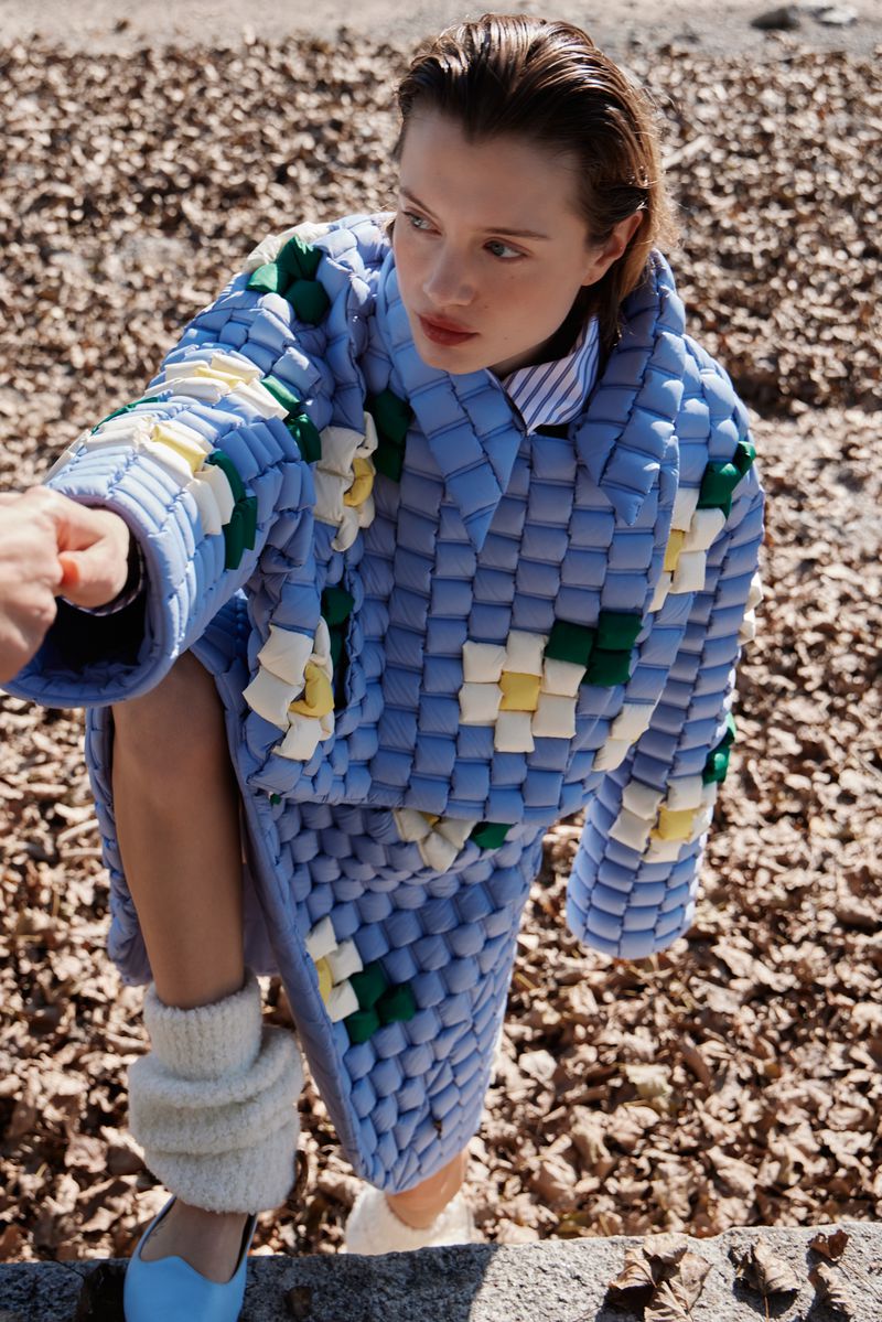 A young woman poses in a distinctive blue and white coat and skirt, standing outdoors with fallen leaves and a hand reaching toward her.