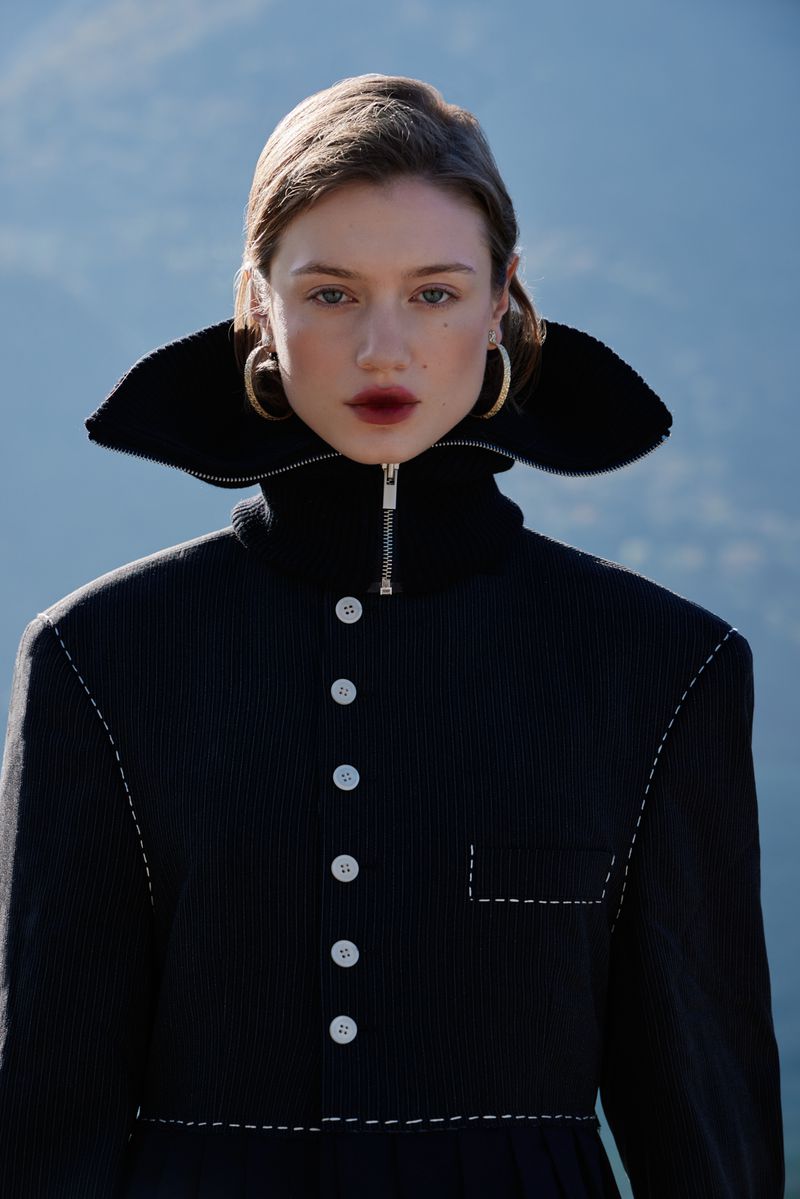 A fashionable young adult female poses in a modern black jacket against a soft blue mountain backdrop, accessorized with gold hoop earrings.