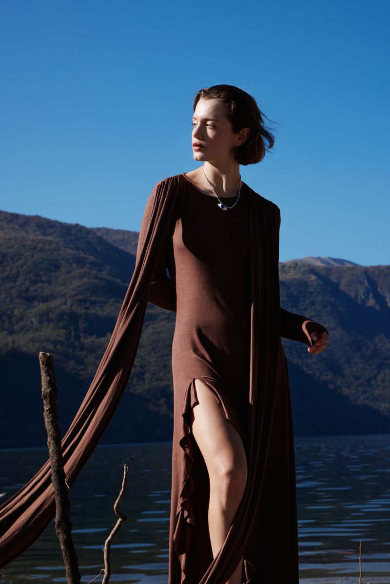 Woman in a flowing brown dress stands against a blue sky, mountain, and lake backdrop on a sunny day in an outdoor setting.