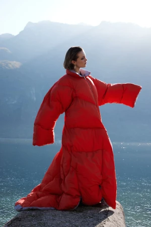 Adult female stands on rock in a bright red oversized puffer coat dress with mountain and lake background during daytime in natural light.