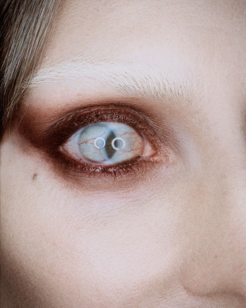 Close-up portrait of a person's face featuring a unique eye with two distinct pupils, dramatic makeup, and light blonde hair.