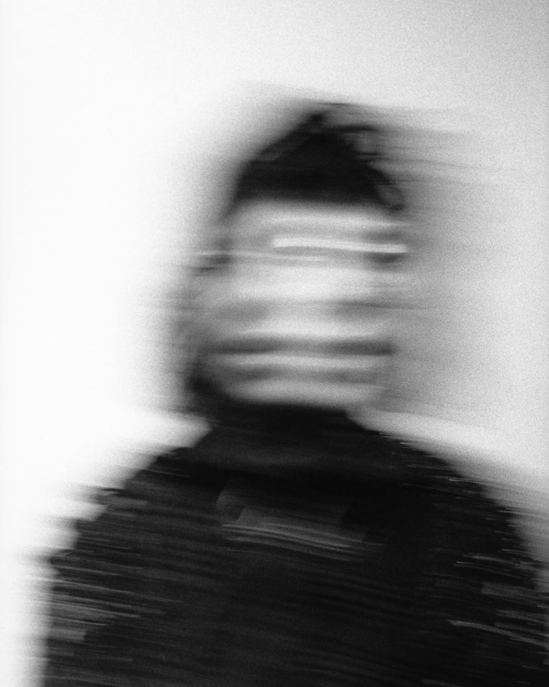 Abstract black and white portrait of an adult with motion blur. The photograph has a grainy texture. The individual has dark hair.
