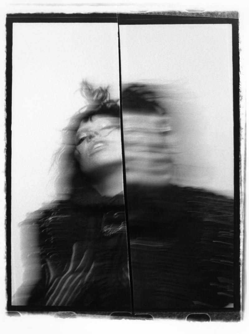Monochromatic motion blur portrait of an adult female with bangs, wearing a dark top, split in two frames, creating a distorted effect.