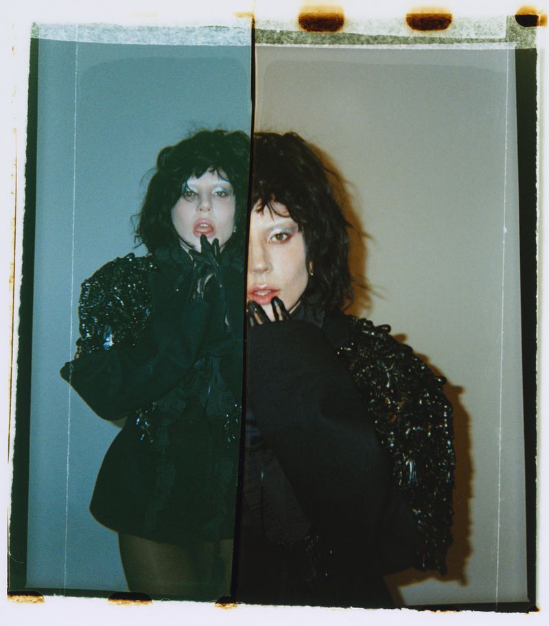 Split-screen portrait of a woman in a black embellished jacket and gloves, with one side having a blue cast in a studio setting.