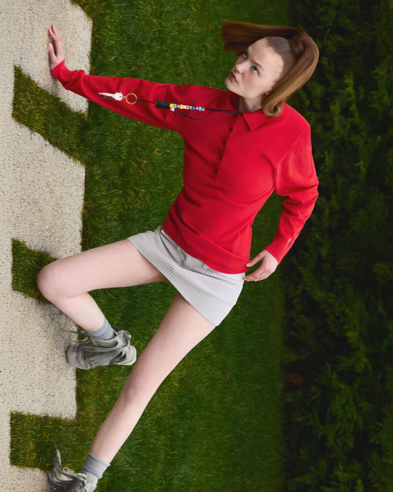 Young woman in a red polo shirt and gray mini skirt poses on a grassy lawn with quirky accessories and sneakers for a fashion shoot.