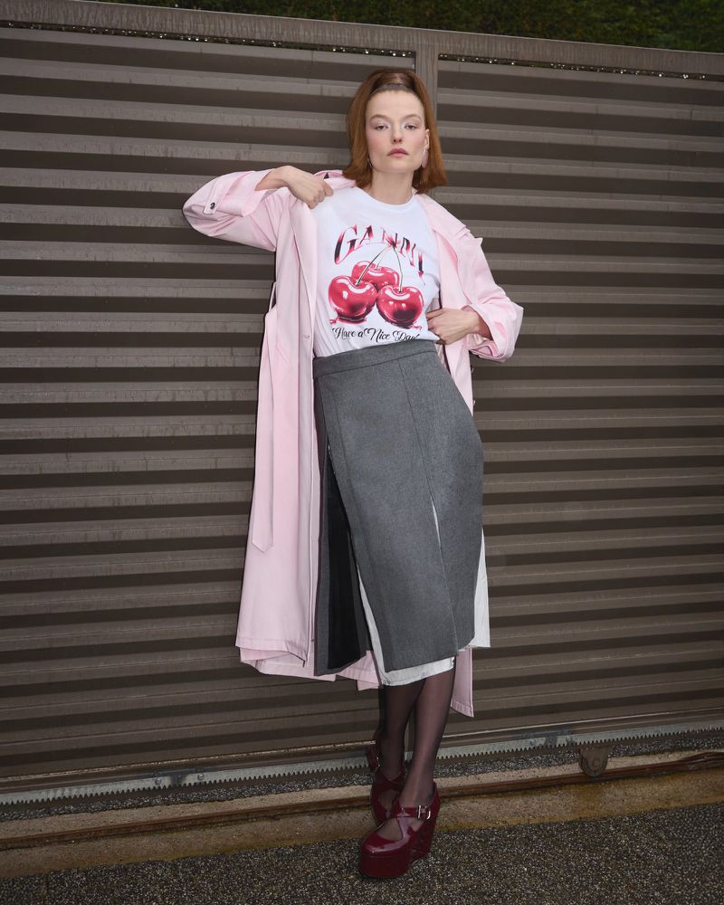 A stylish woman poses in a pink coat, cherry-print tee, and gray skirt against a metal wall. She accessorizes with platform shoes.
