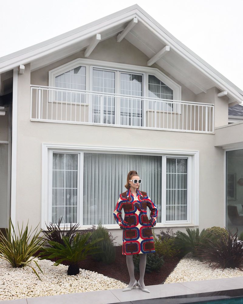 Fashionable woman in patterned coat and sunglasses stands confidently against a modern house backdrop, embodying retro chic style.