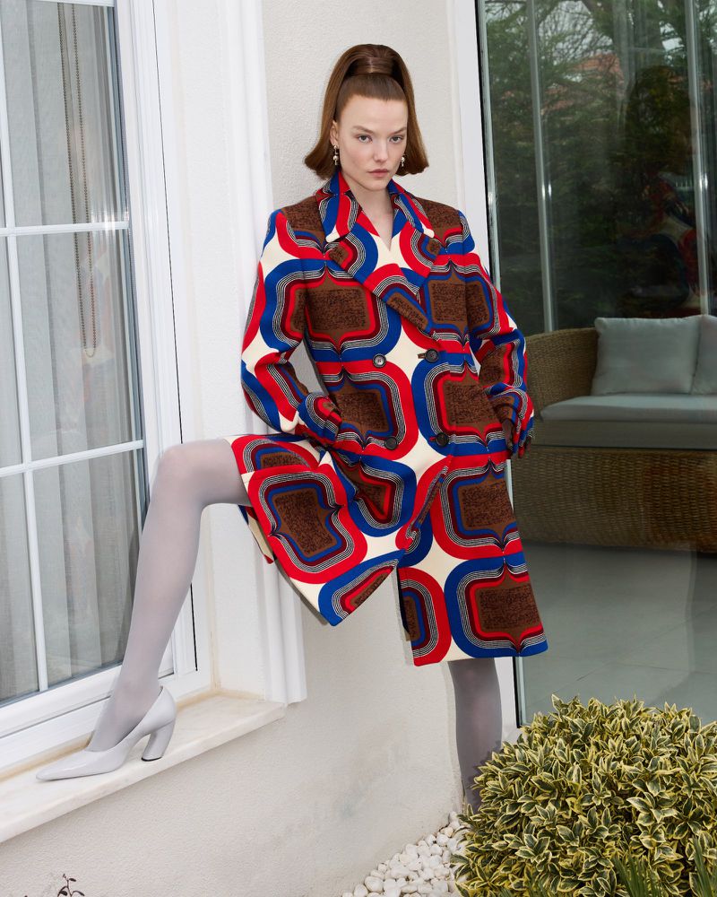 A female model poses in a patterned coat and gray tights while leaning against a white wall with an outdoor backdrop, indoor setting.