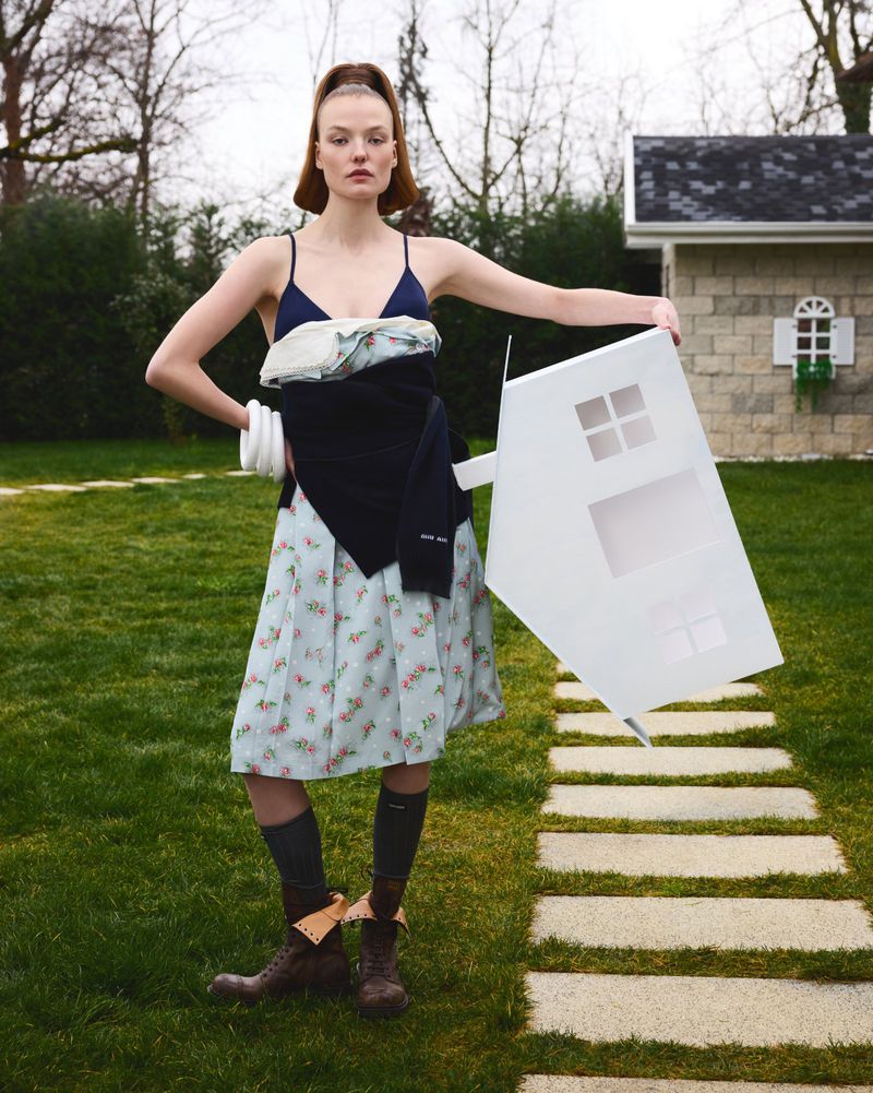Adult female model in light blue dress holds white toy house outdoors on grass with small outbuilding. Fashion editorial image.